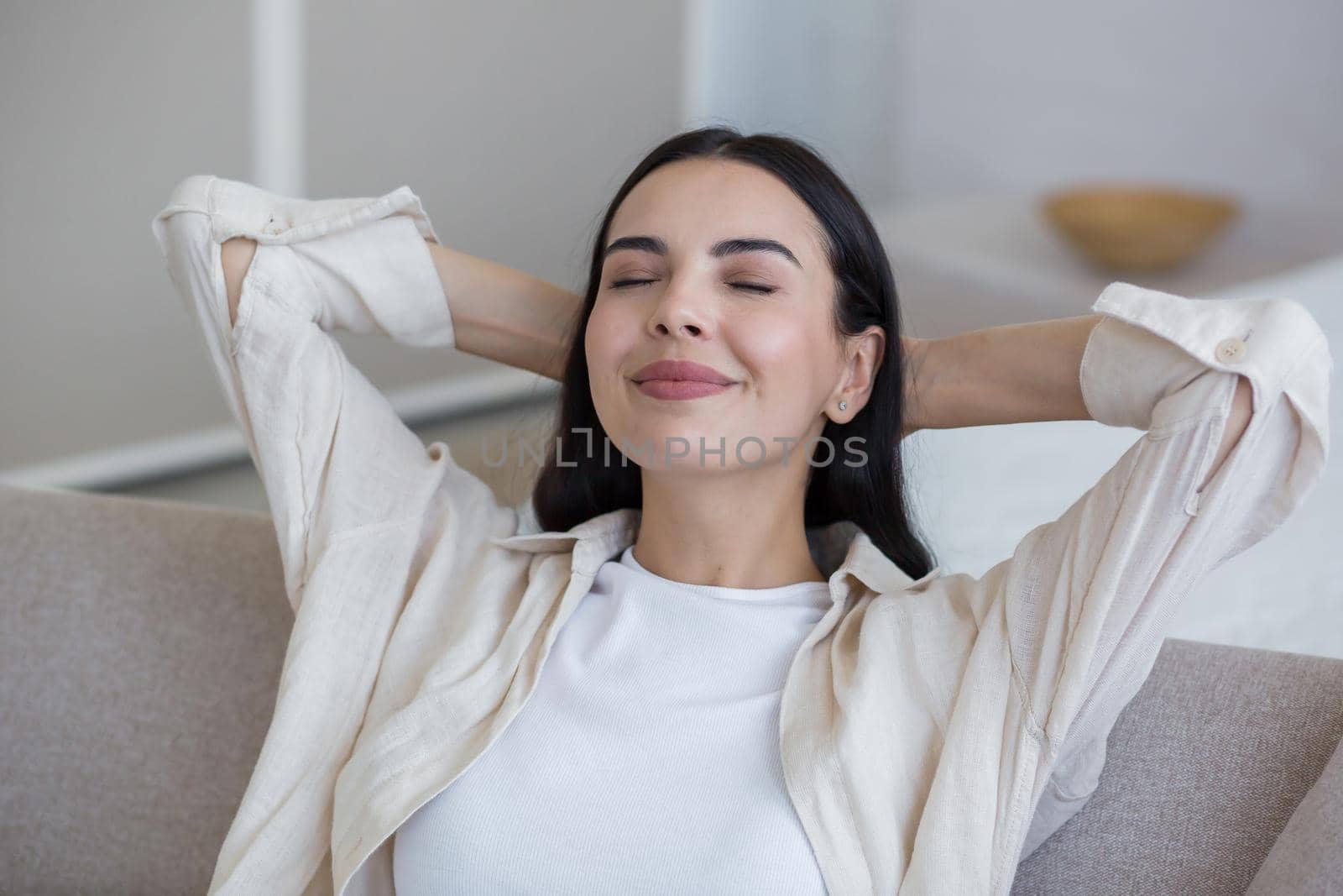 Close up photo. Happy young beautiful woman relaxing at home on sofa with eyes closed, arms behind head. Enjoys silence, peace, dreams, smiles. Have a nice weekend.