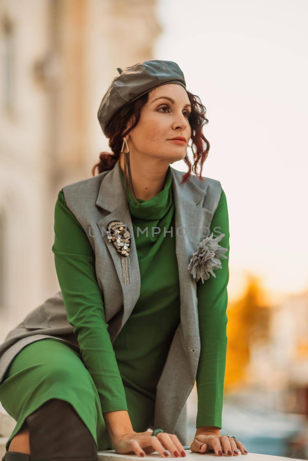 Outdoor fashion portrait of an elegant fashionable brunette model in a beret, green dress and a gray waistcoat posing at sunset in a european city in autumn