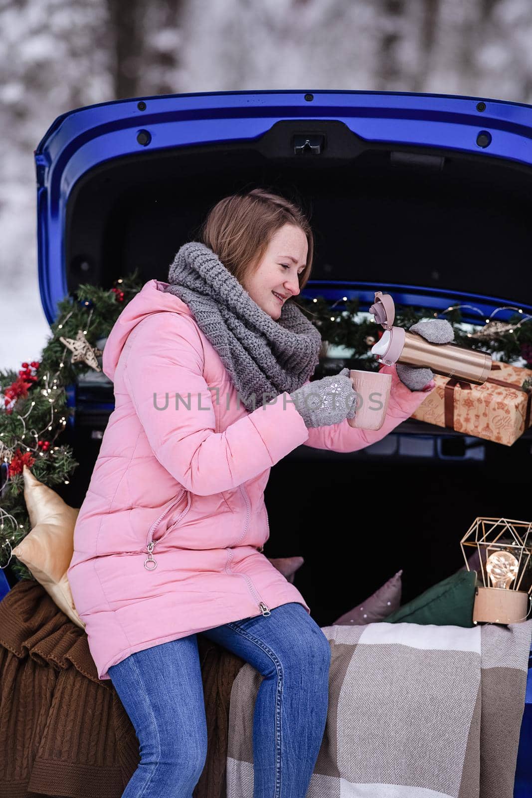 A woman in winter clothes drinks a hot drink sitting in a car decorated in a New Year's way. Traveling by car through the forest. A trip before Christmas.