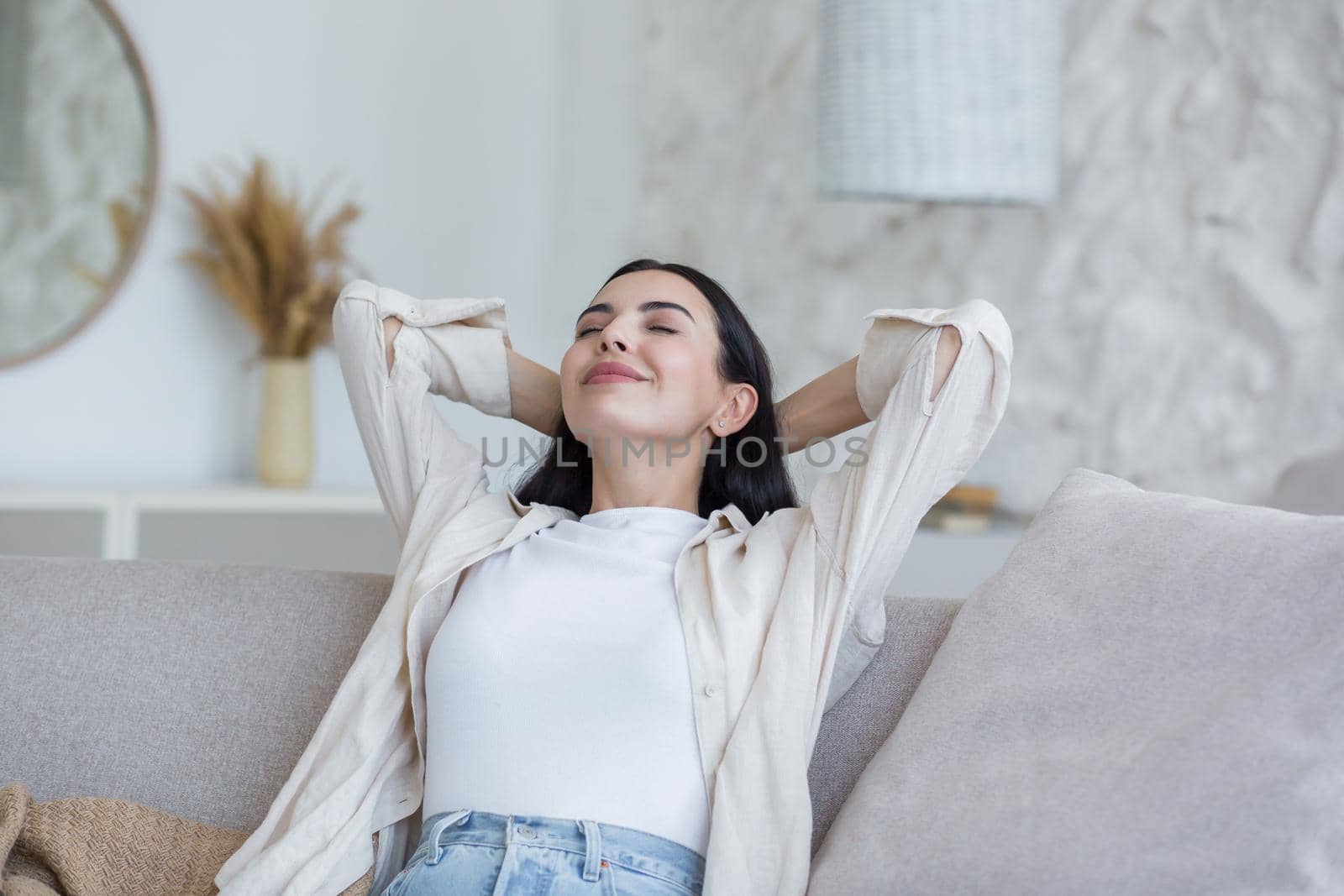 Portrait happy young beautiful woman relaxing at home on sofa with eyes closed, arms behind head. Enjoys silence, peace, dreams, smiles. Have a nice weekend.