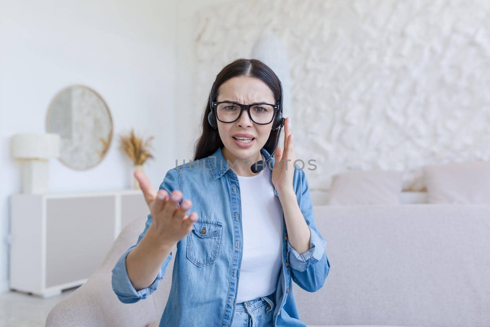 Webinar. Young beautiful business woman in glasses and headphones sits on the sofa at home and conducts a webinar, online training on camera, consults, tells, gestures with hands, smiles.