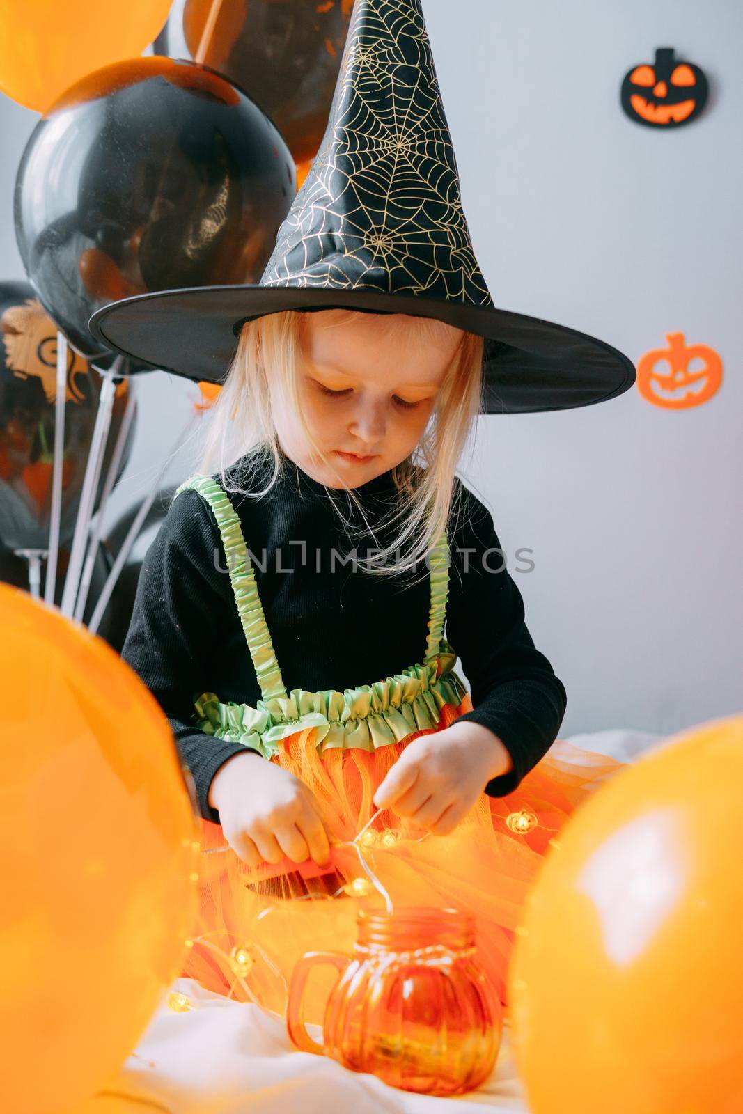 Children's Halloween - a girl in a witch hat and a carnival costume with airy orange and black balloons at home. Ready to celebrate Halloween.