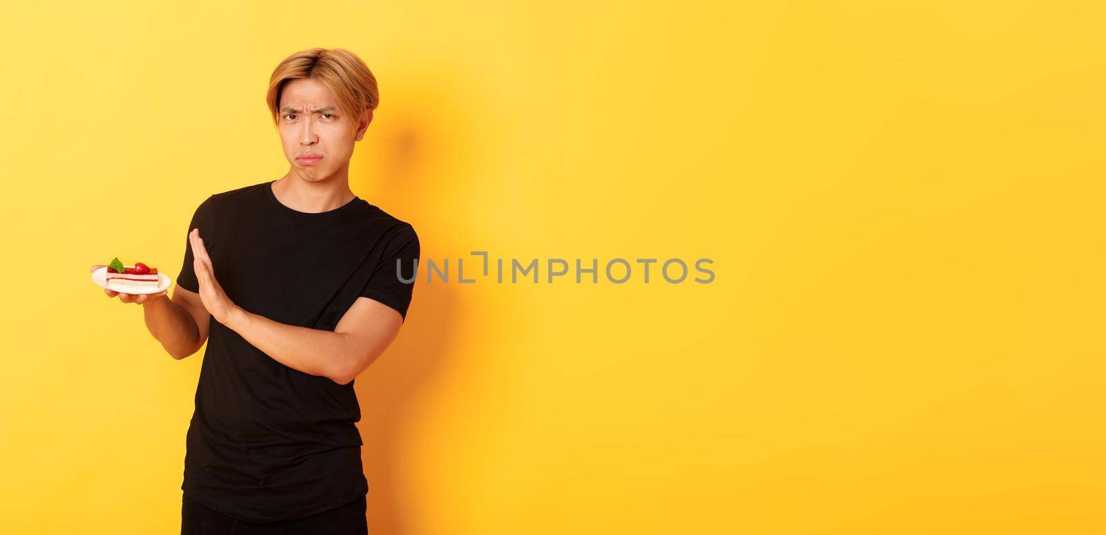 Portrait of disgusted and upset young asian guy refuse eating cake, looking displeased and showing rejection gesture, yellow background.