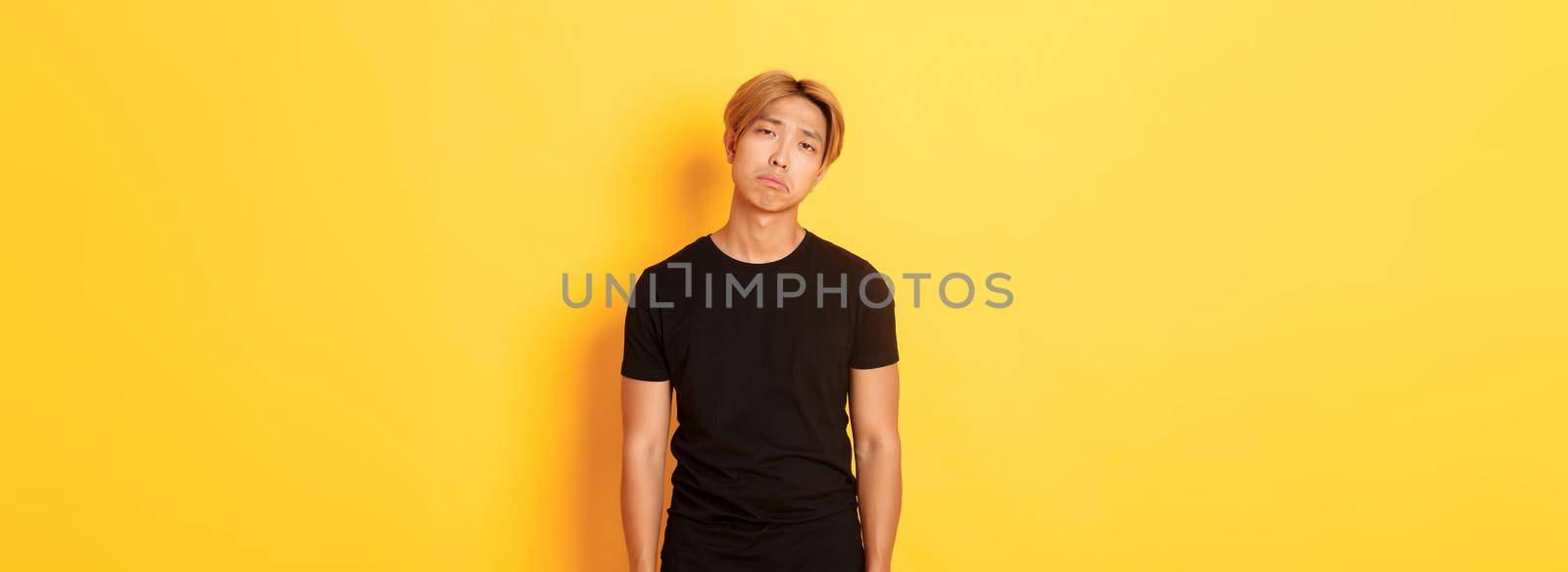 Portrait of exhausted and sad asian guy looking sad, standing drained over yellow background.