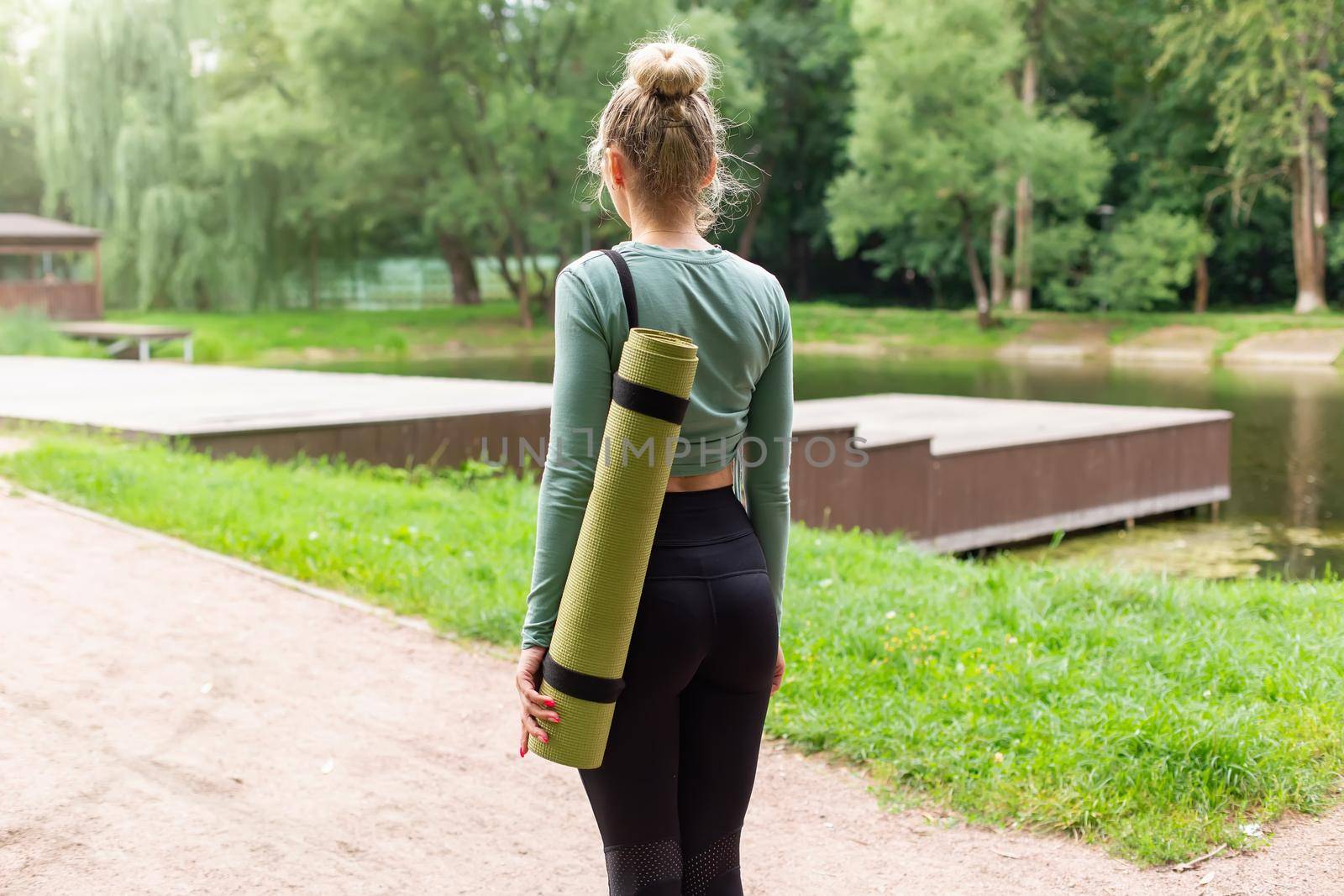 Rear view, slender blonde woman in the park in summer, with a green gym mat behind her back. Close up. Copy space