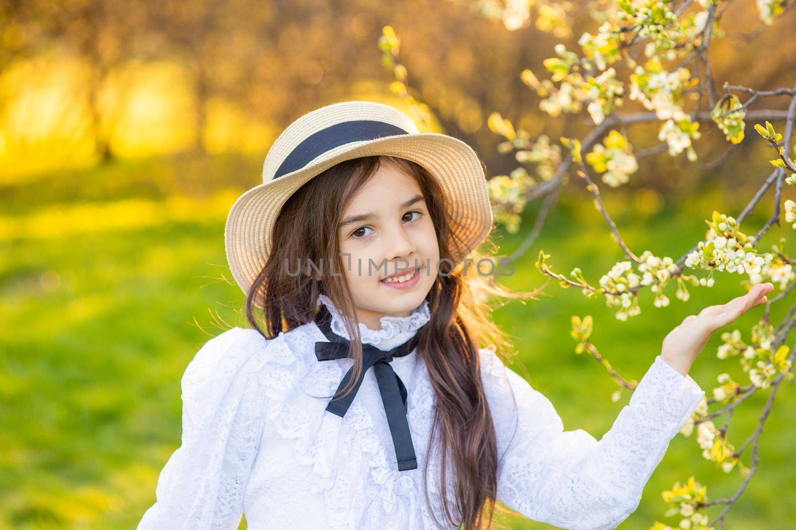 a smiling adorable girl in a hat and white openwork dress , holding a branch of a tree blooming with white flowers, in the spring in the park. Close up