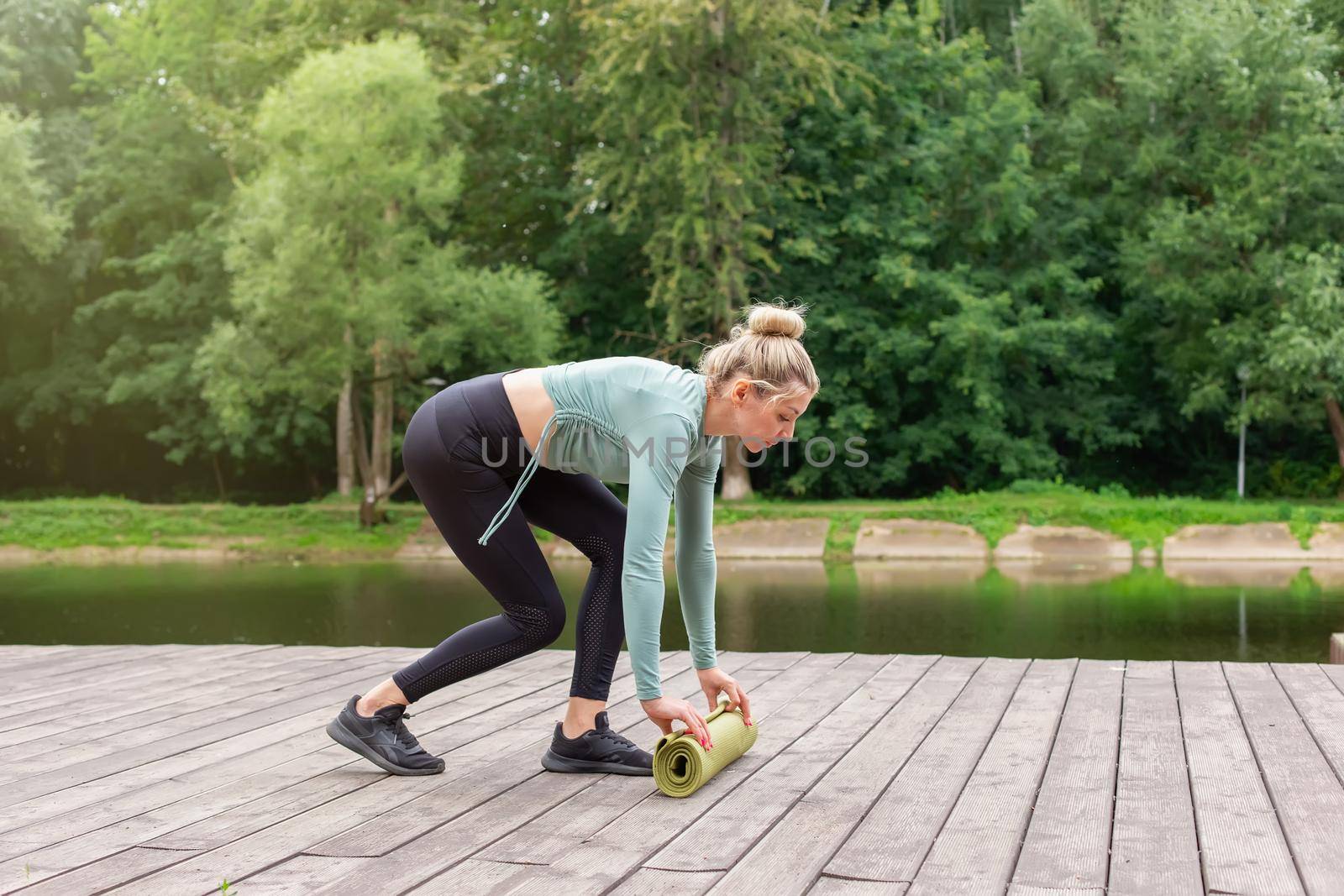 A woman in a green top and black leggings, in the summer in the park, unwinds a green gym mat, prepares for sports. side view. Copy space