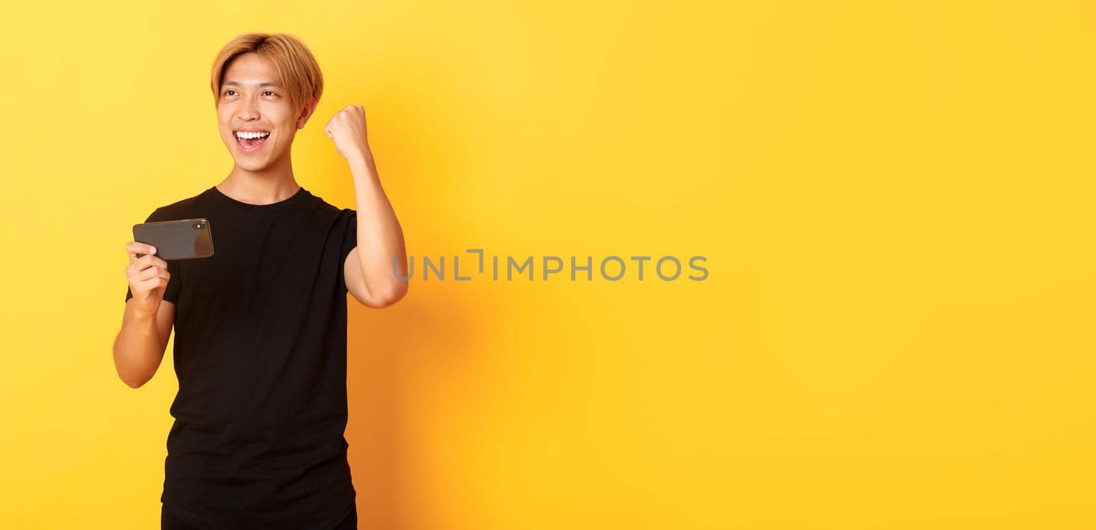 Portrait of stylish handsome asian guy playing smartphone game, rejoicing from winning, standing yellow background.