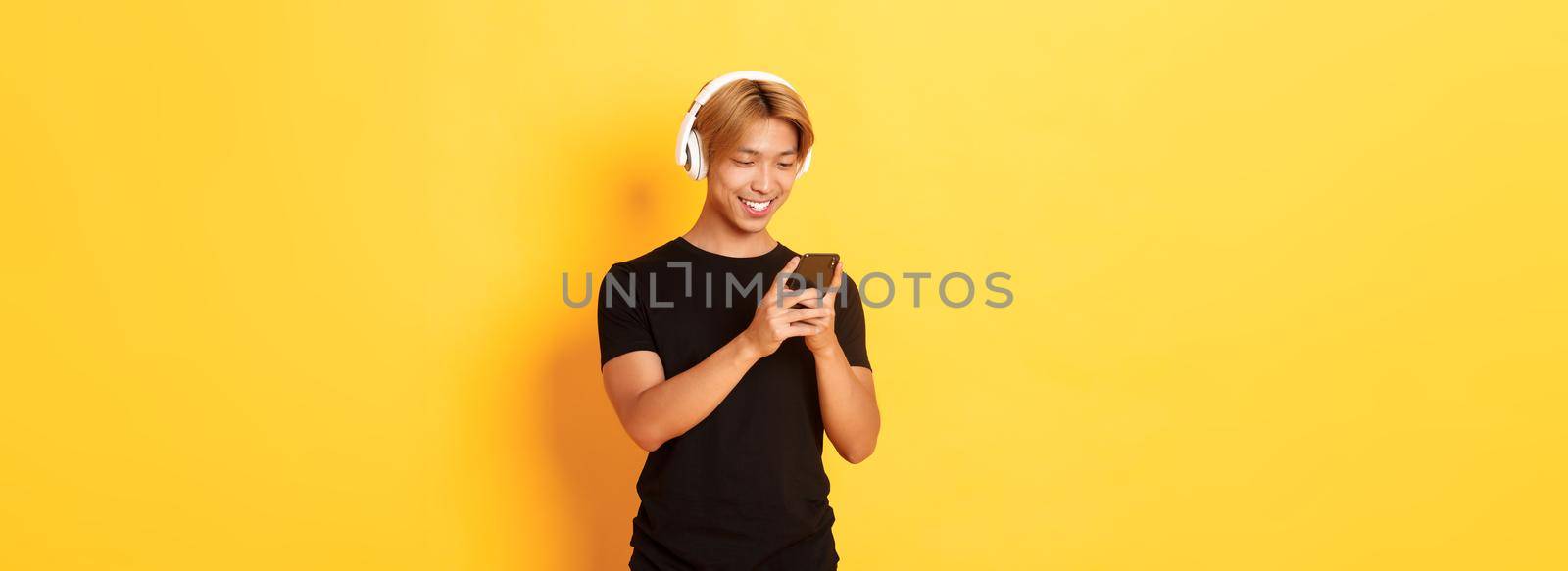 Young smiling asian guy using smartphone and listening music or podcast in wireless headphones, standing yellow background by Benzoix