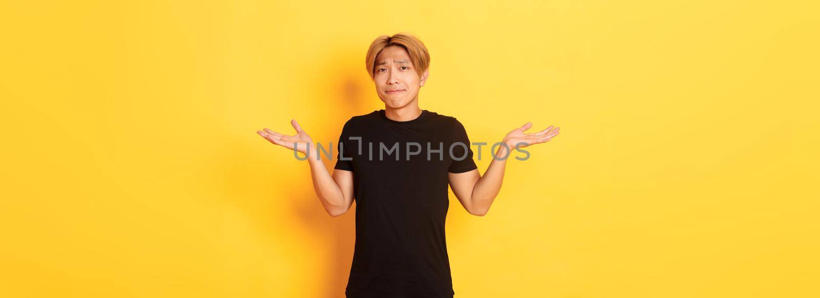 Portrait of confused and indecisive attractive korean guy, shrugging and pouting upset, standing yellow background by Benzoix