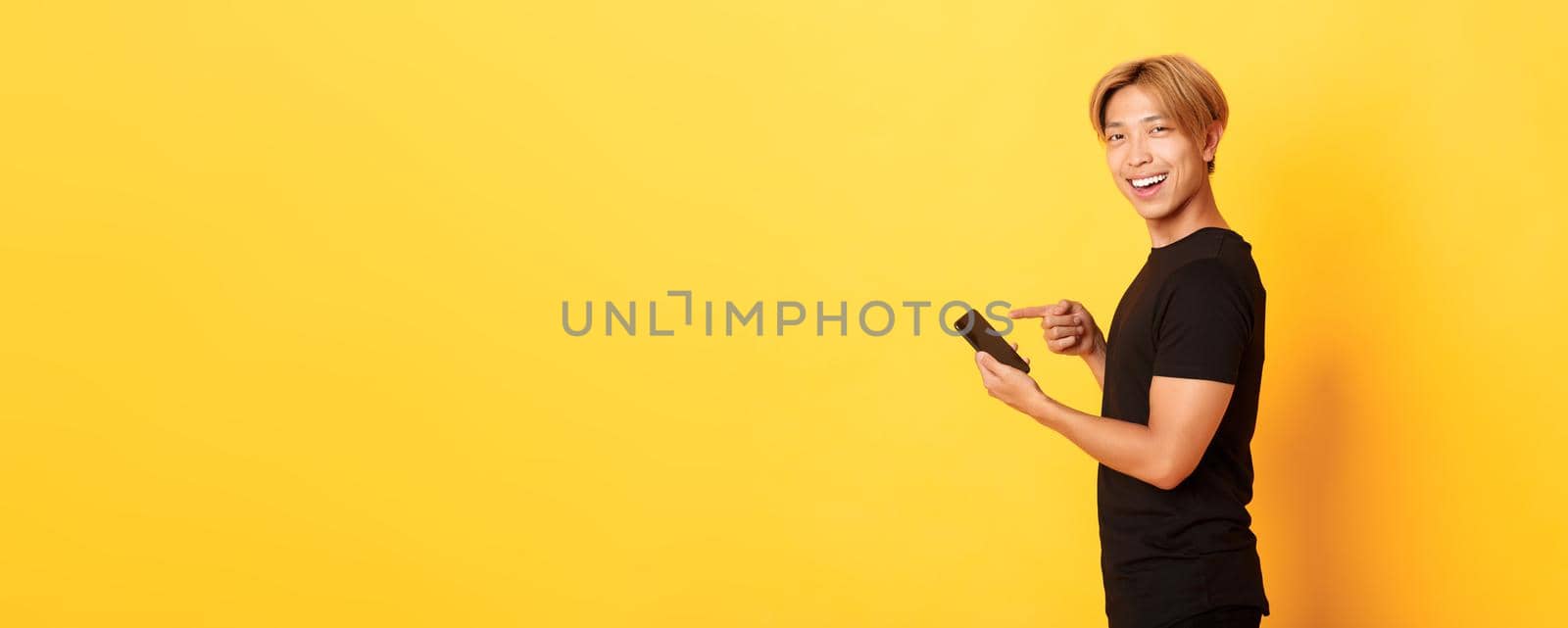 Portrait of pleased smiling handsome asian man, standing in profile and pointing finger at smartphone, recommend app, standing yellow background by Benzoix
