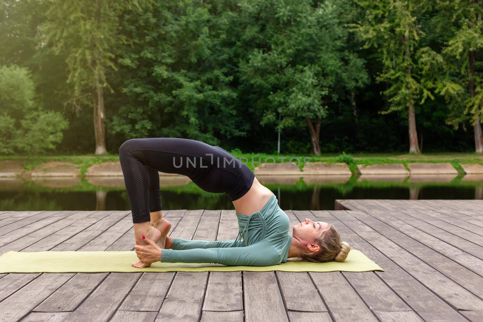 A woman on a wooden platform by a pond in a park in summer, does yoga, performing a table pose. Copy space