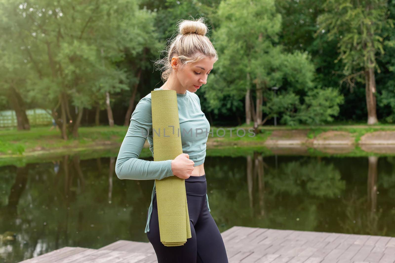 A beautiful slender blonde, holding a green gym mat in her hands, stands in the park by the pond in summer, looking down. Copy space