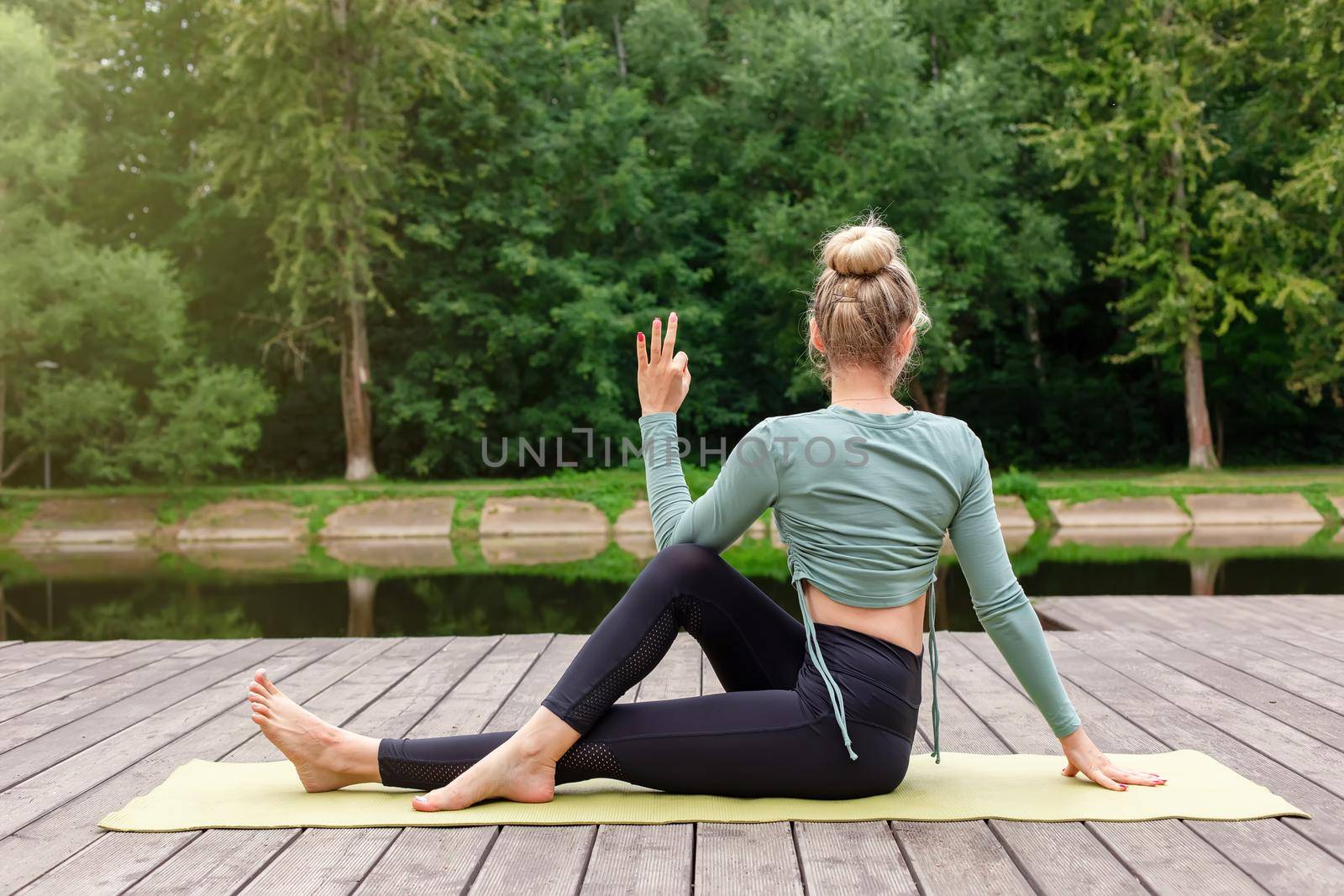 A woman in a green top and leggings, sits on a wooden platform by a pond in a park in summer, does yoga, performing a table pose. Copy space. Back view