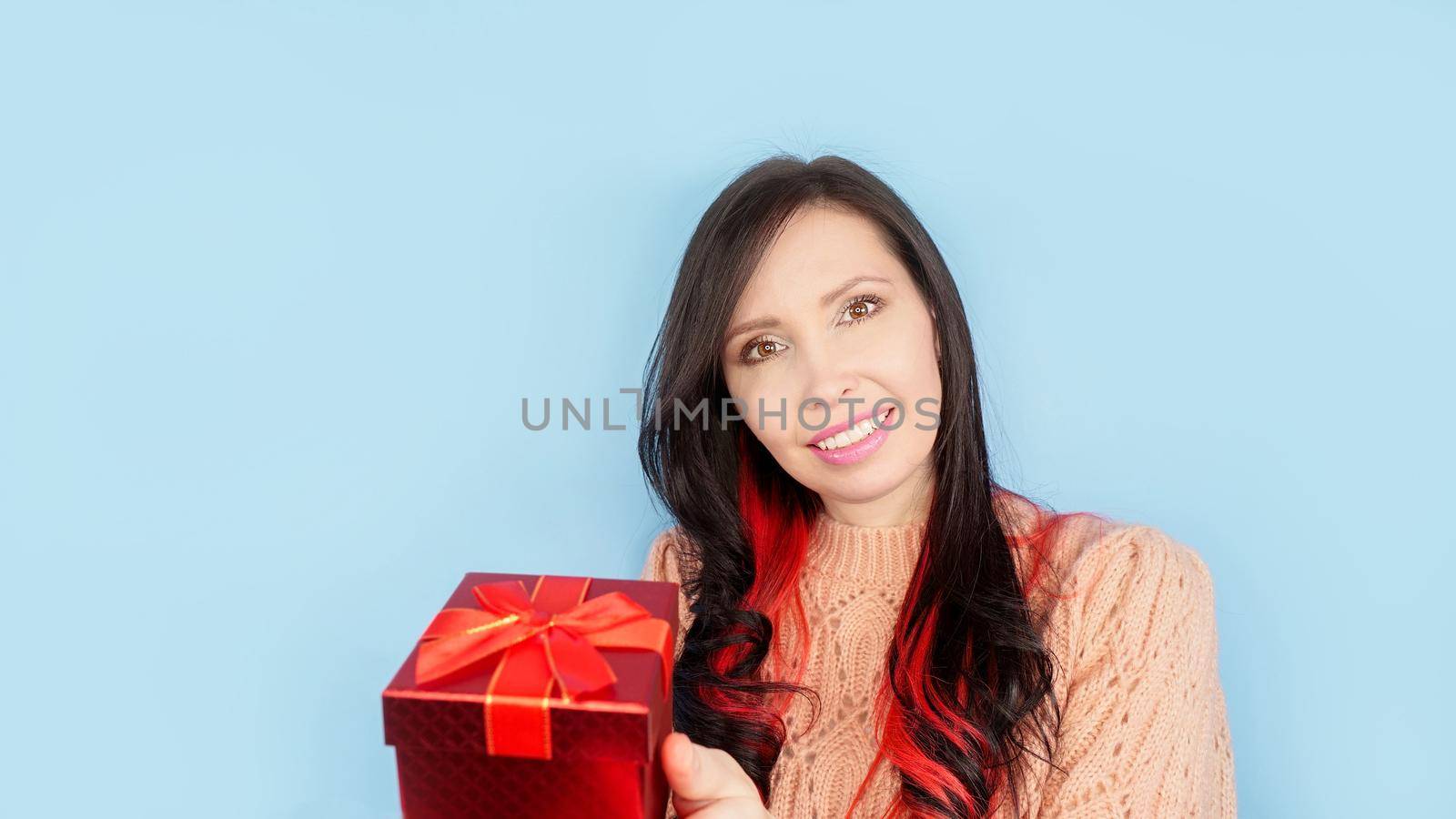 Photo of pretty excited lady curly brunette with red hair smiling hold red gift box wear coral pullover isolated blue color background. Christmas holidays, Birthday.