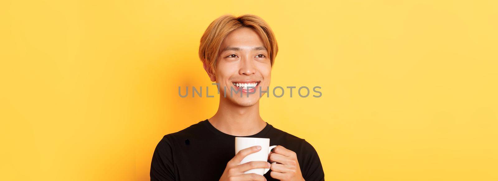 Close-up of happy smiling handsome blond asian guy, looking away with dreamy nostalgic look while drinking coffee, standing over yellow background.