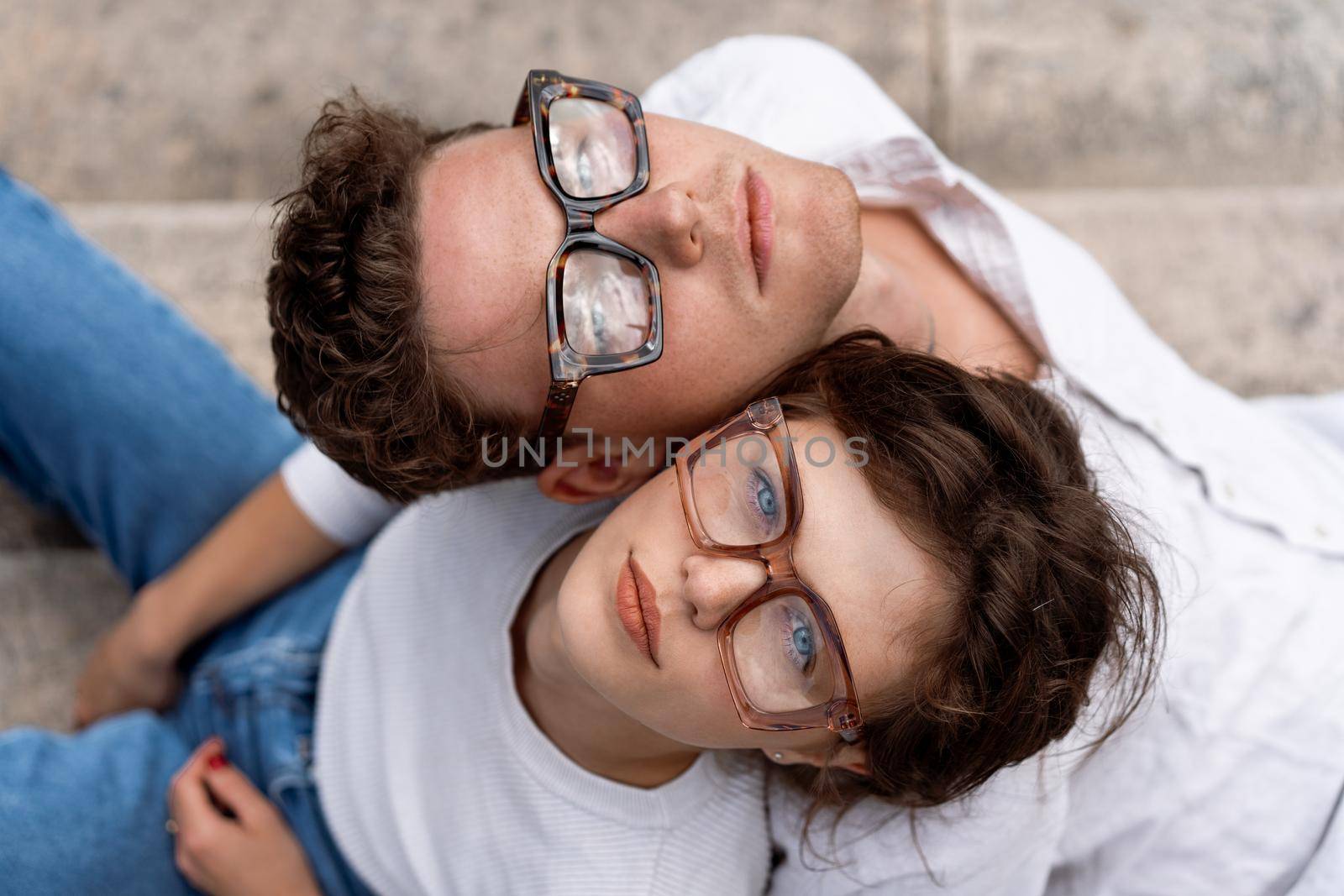 Portrait of two attractive students man and woman wearing stylish glasses sitting floor outdoor and smiling, head to head cheek to cheek, break relax between lesson, work, projects Eyeglasses concept