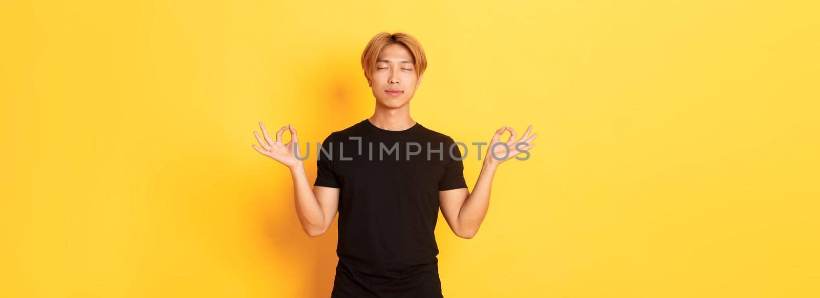 Portrait of relaxed and peaceful handsome asian man with blond hair, close eyes and meditating, attend yoga, standing yellow background.