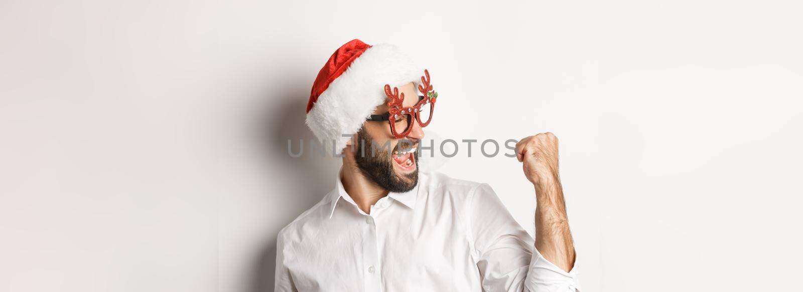 Close-up of happy bearded man wearing santa hat and christmas party glasses, making fist pump and rejoicing, achieve goal and celebrating, white background by Benzoix