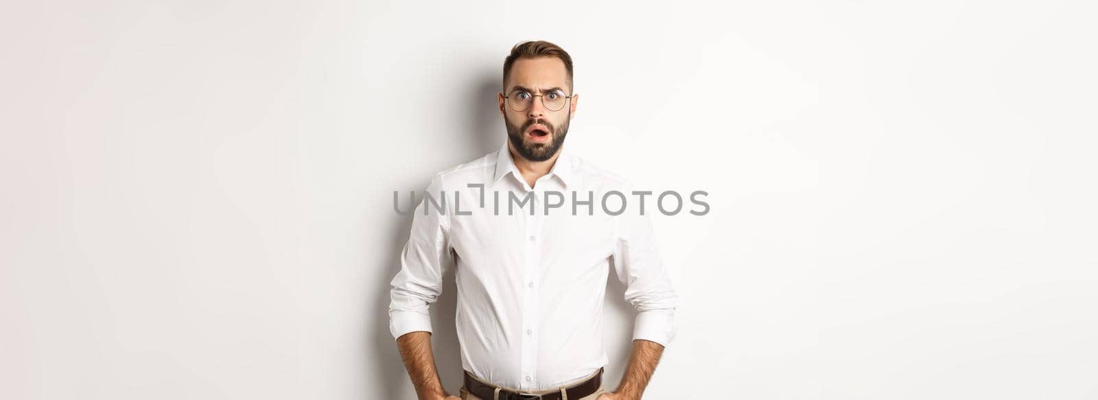 Shocked and displeased businessman in glasses, gasping and looking upset at camera, standing over white background. Copy space