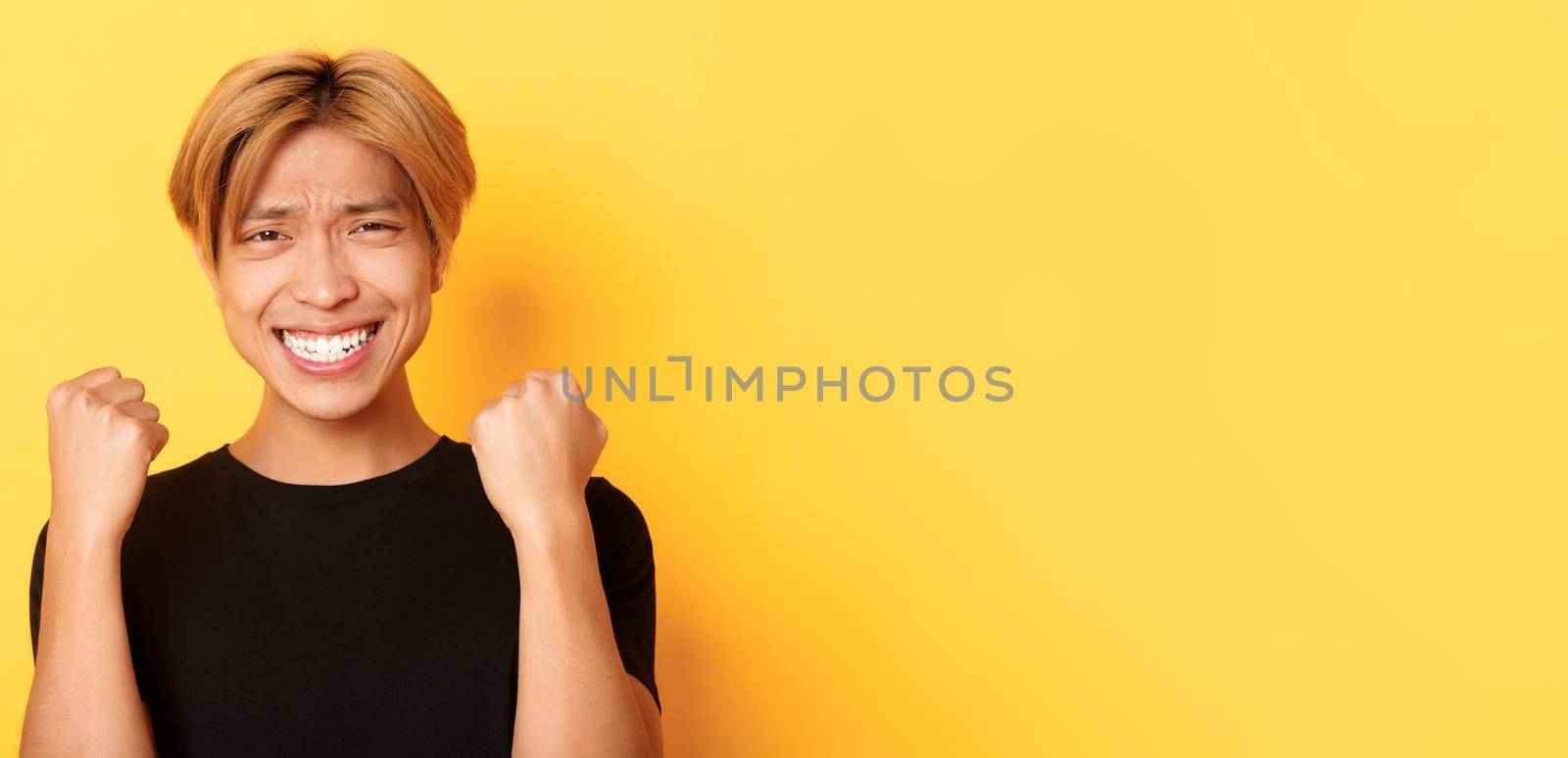 Close-up of lucky happy asian guy winning prize, making fist pump gesture and smiling satisfied, triumphing over achievement, standing yellow background by Benzoix