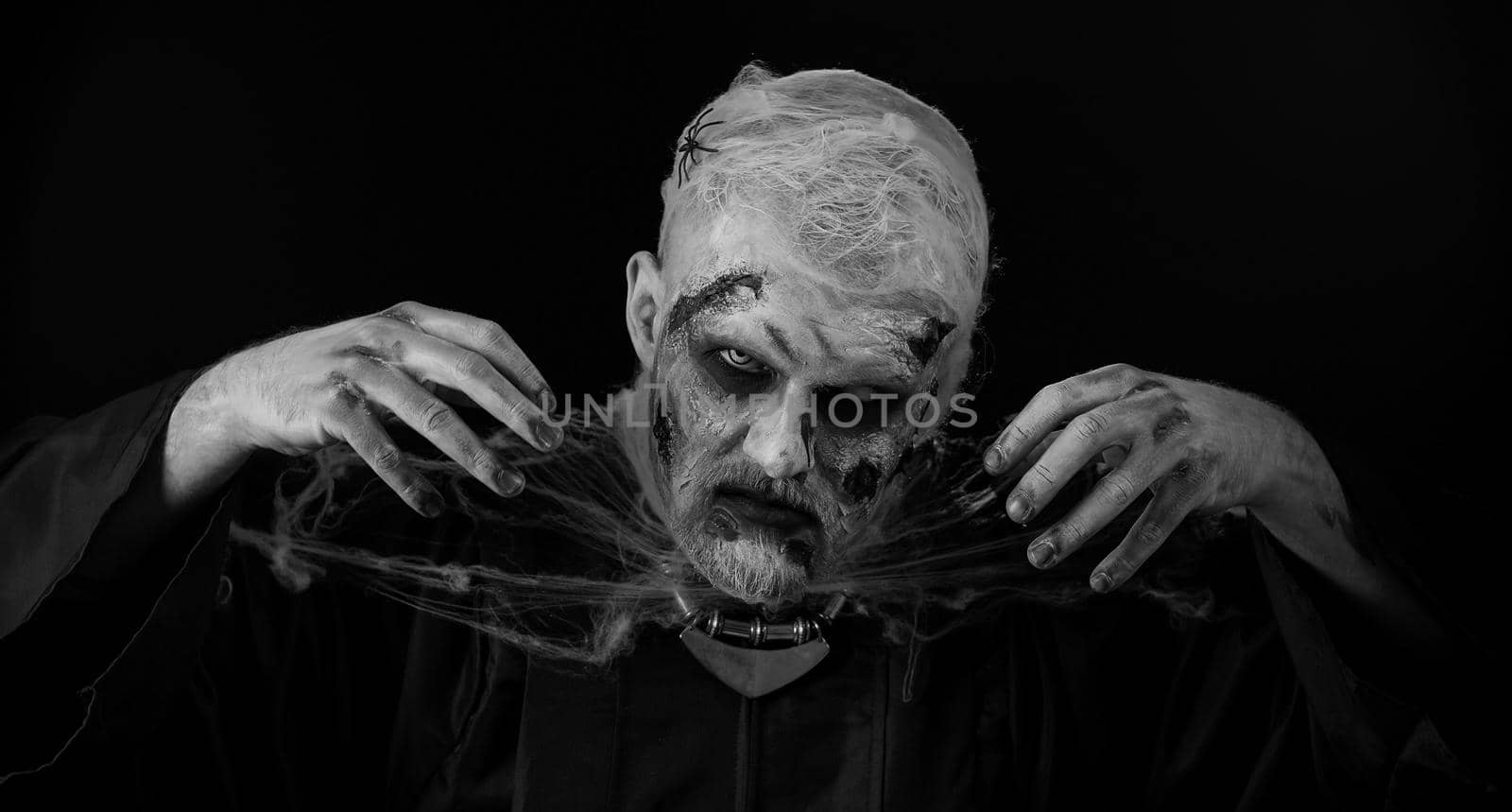 Sinister man with horrible scary Halloween zombie makeup in costume making faces, looking ominous at camera, trying to scare. Dead guy with wounded bloody scars face isolated against black background