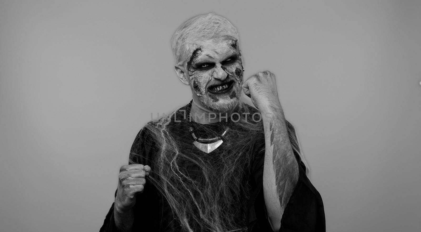 Spooky crazy zombie man alone with makeup with fake wounds scars celebrate success win scream rejoices doing winner hands gesture say Yes in gray studio. Sinister dead guy. Halloween holidays