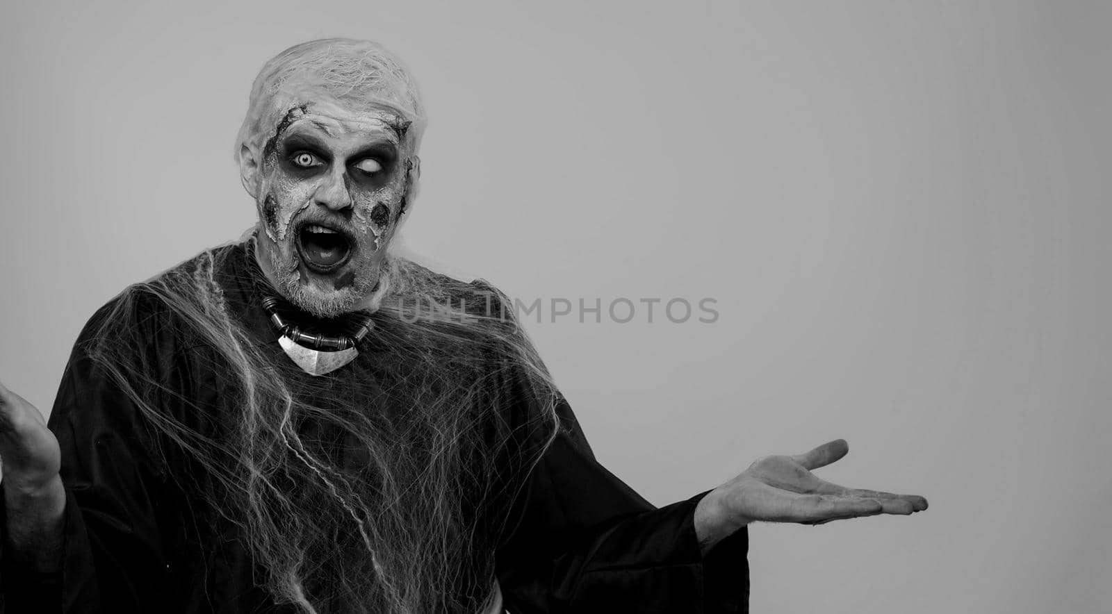 Creepy man with bloody scars face, Halloween stylish zombie make-up. Scary wounded undead guy showing thumbs up and pointing at left on blank space. Place for your advertisement logo. Sinister monster