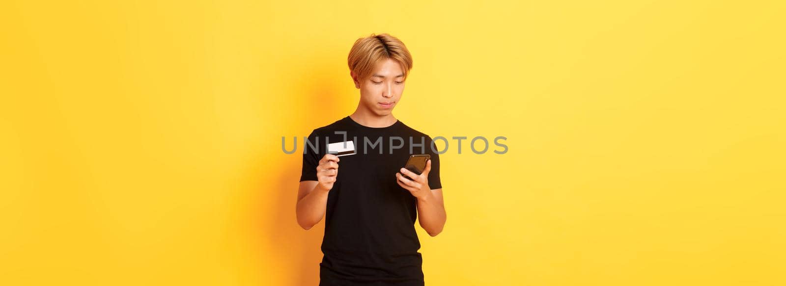 Young handsome asian guy in black t-shirt shopping online, holding credit card and using smartphone, standing yellow background.