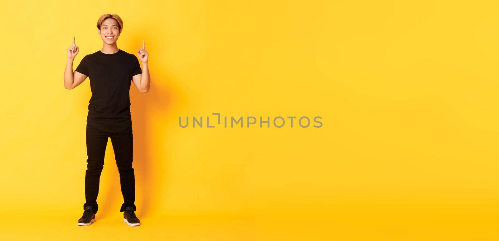 Full-length of confident handsome asian man with blond hair, smiling and pointing fingers up, showing logo, yellow background.