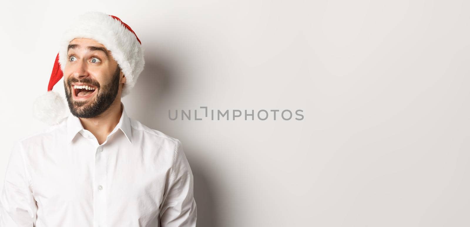 Close-up of amazed bearded man in santa hat, looking left with excitement, christmas promo offer, standing over white background.