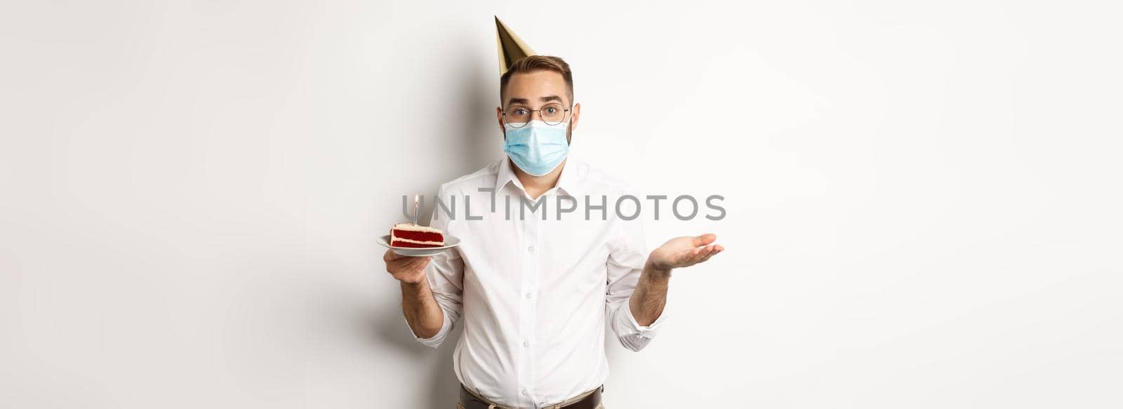 Coronavirus, quarantine and holidays. Confused man in face mask, holding birthday cake and shrugging, standing over white background clueless.