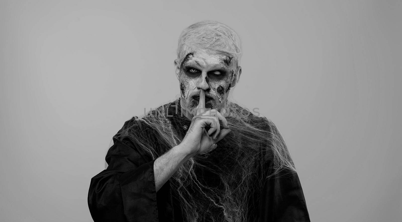 Shh be quiet. Sinister man with horrible scary Halloween zombie makeup in costume presses index finger to lips makes silence gesture sign do not tells secret. Dead guy with wounded bloody scars face