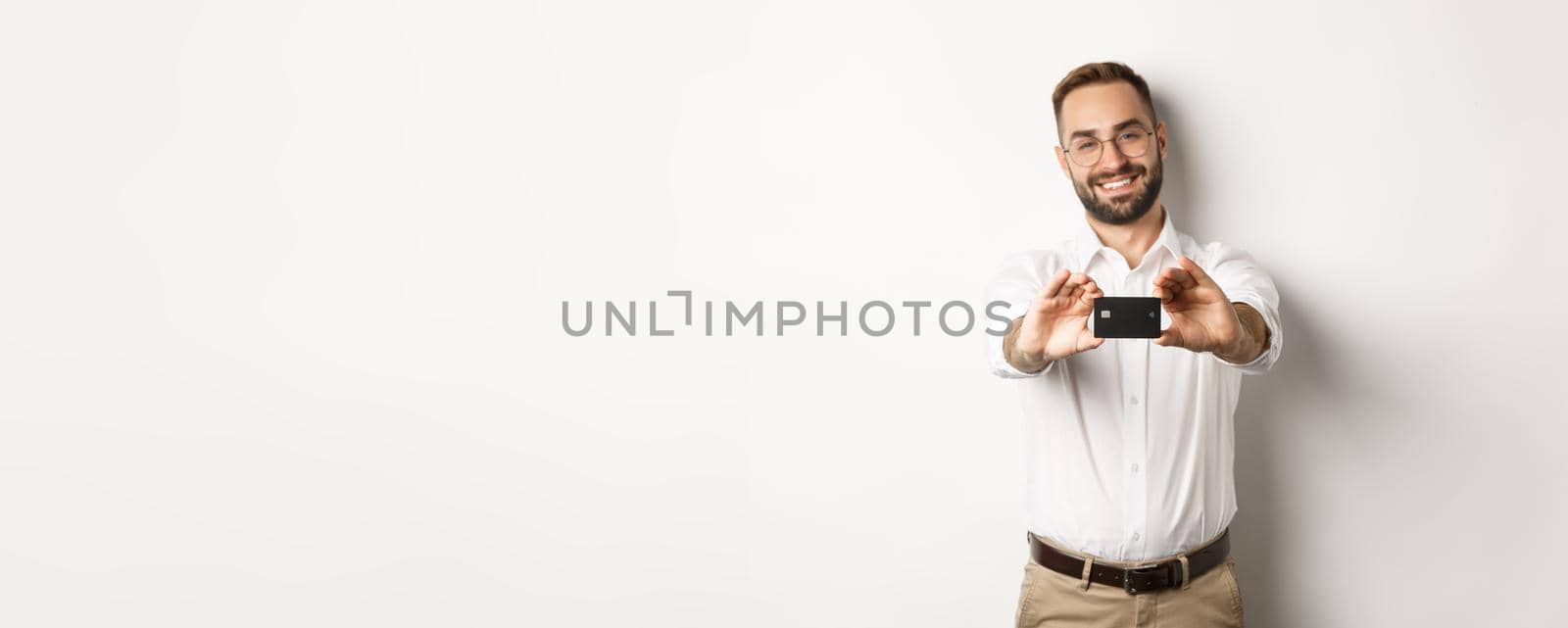 Handsome man in glasses holding a credit card, smiling pleased, standing over white background.