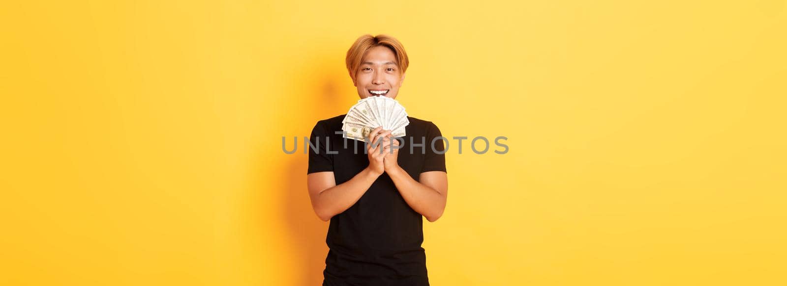 Cheerful winning handsome asian guy, smiling pleased and holding money, feeling lucky, standing yellow background by Benzoix