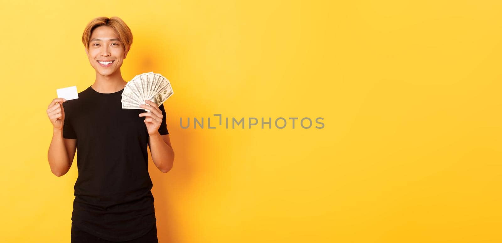 Pleased handsome asian guy showing money and credit card, smiling happy, standing yellow background.