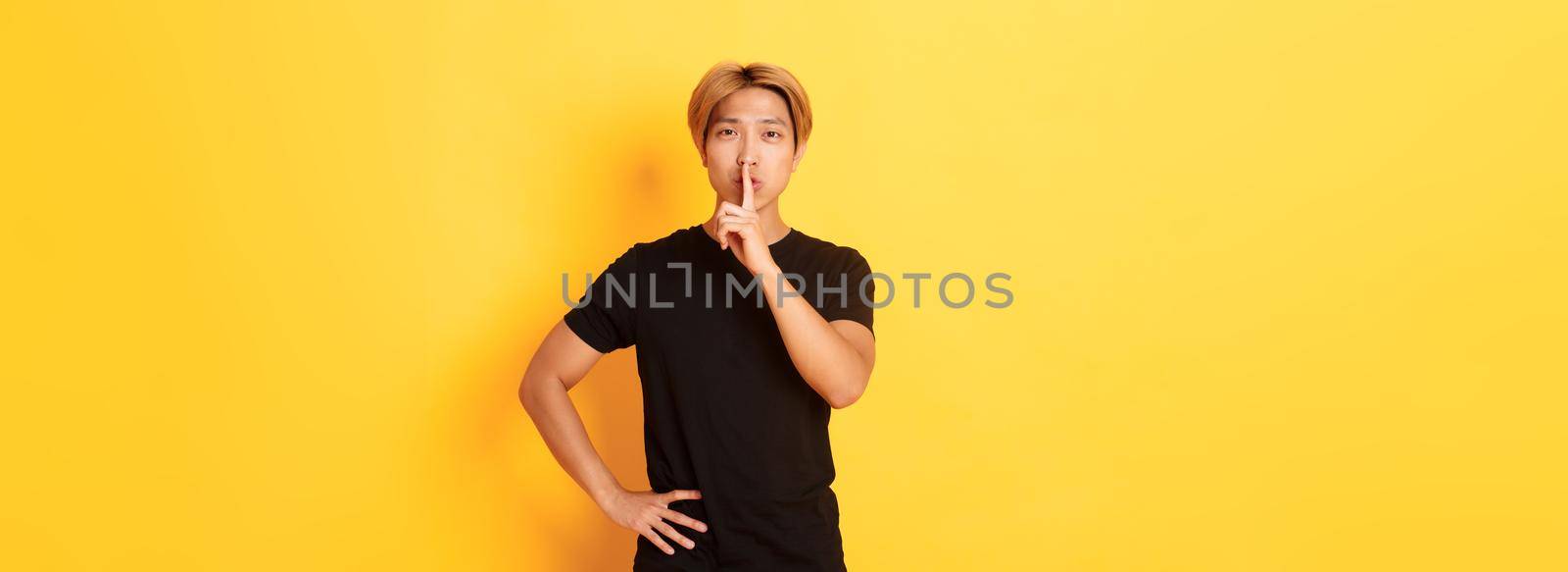 Portrait of attractive blond asian man shushing, press finger to lips and hush, yellow background.