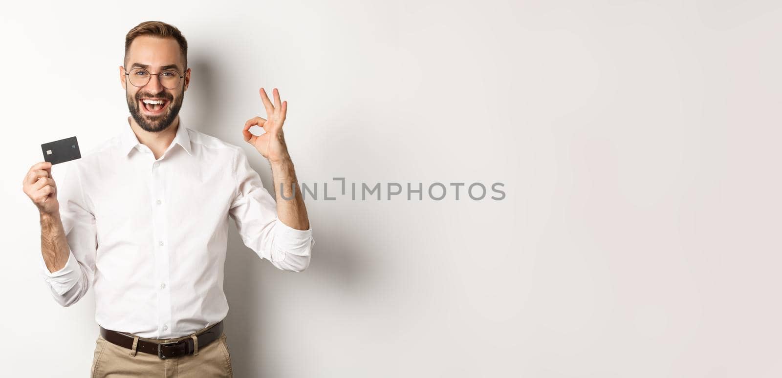 Handsome man showing his credit card and okay sign, recommending bank, standing over white background by Benzoix