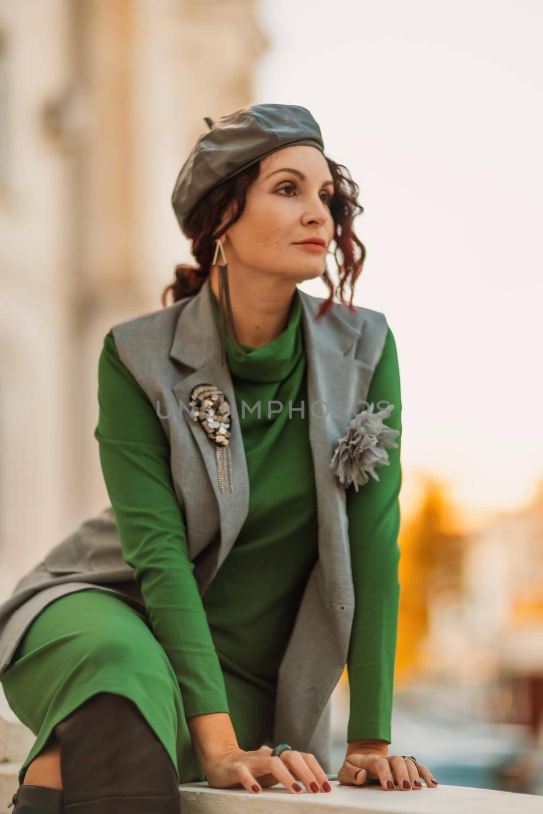 Outdoor fashion portrait of an elegant fashionable brunette model in a beret, green dress and a gray waistcoat posing at sunset in a european city in autumn