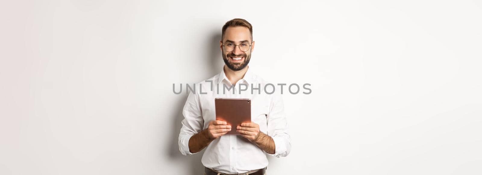 Handsome bearded man in glasses using digital tablet, smiling satisfied, standing against white background.