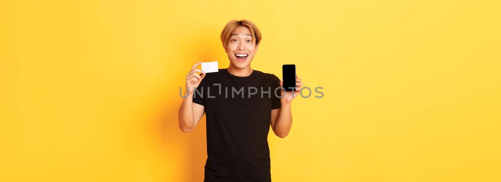 Portrait of excited happy asian man showing mobile phone screen and credit card with joyful smile, standing yellow background by Benzoix