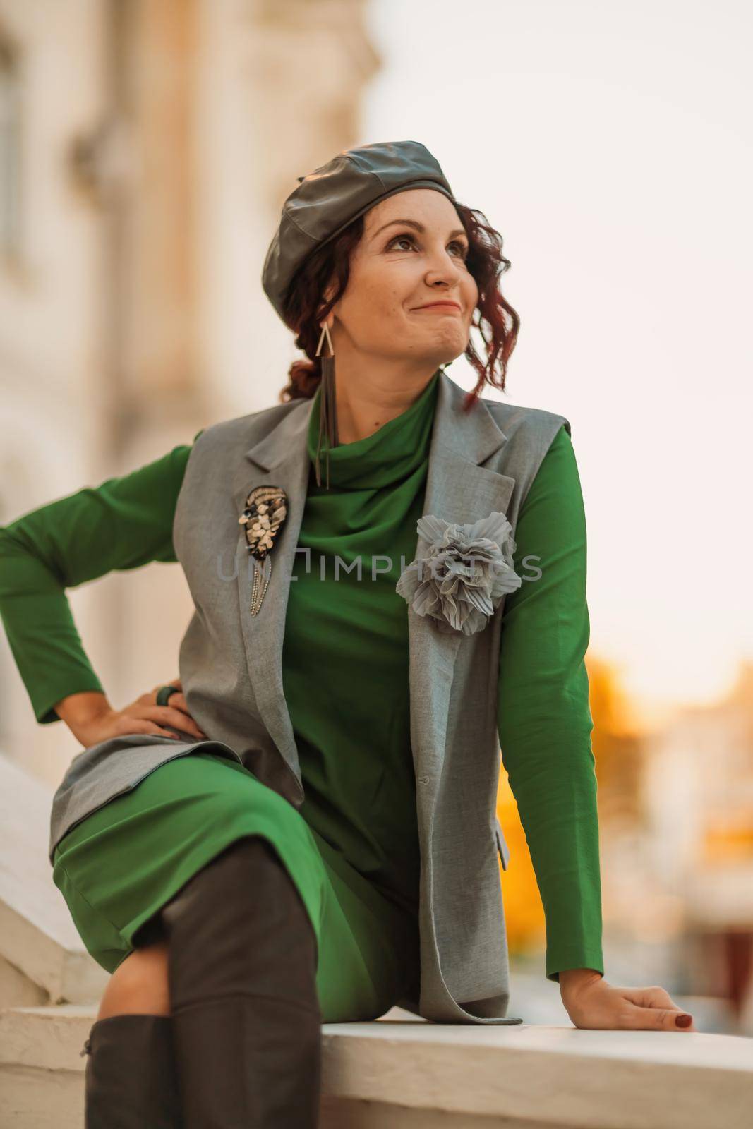 Outdoor fashion portrait of an elegant fashionable brunette model in a beret, green dress and a gray waistcoat posing at sunset in a european city in autumn