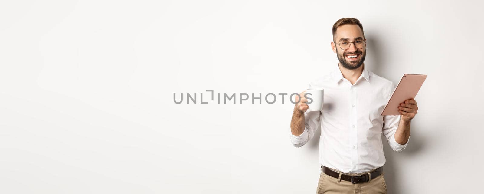 Satisfied boss drinking tea and using digital tablet, reading or working, standing over white background.