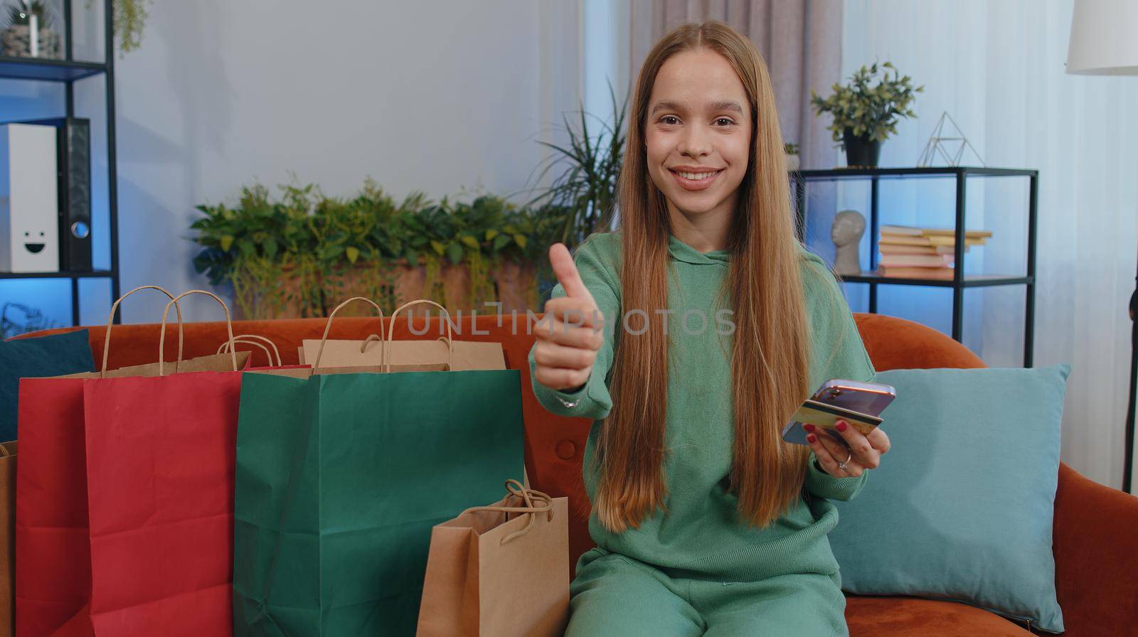 Happy shopaholic consumer girl sits with shopping bags at modern home apartment making online order, payment with credit plastic bank card. Young child kid on couch at home. Discount sale on internet
