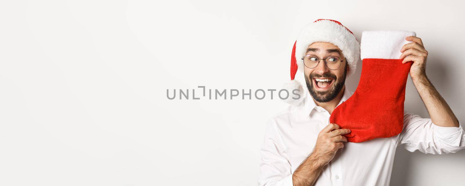 Close-up of happy man celebrating christmas, receive gifts in xmas sock and looking excited, wearing santa hat and glasses, white background.