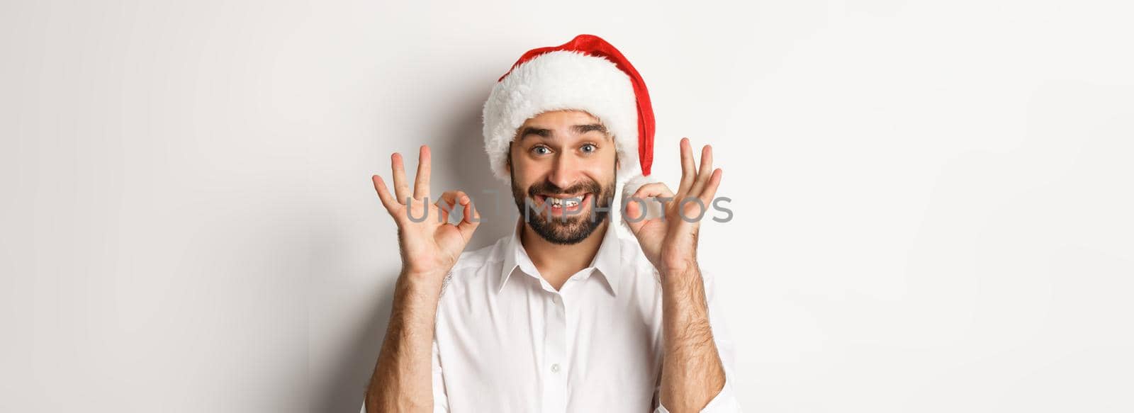 Party, winter holidays and celebration concept. Joyful man enjoying christmas and showing okay sign, smiling satisfied, wearing santa hat, white background.
