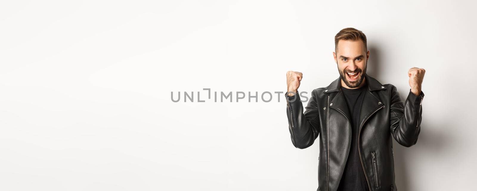 Confident and assertive man in leather jacket making fist pump, rejoicing of winning, feeling encouraged and satisfied, white background.
