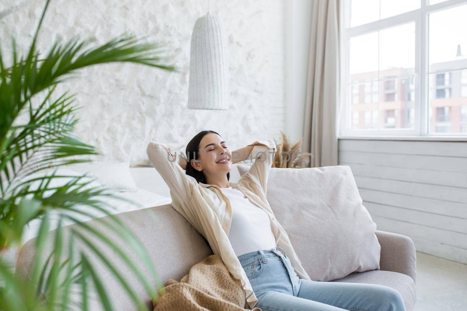 A young beautiful business woman is resting on the sofa after a successful working day. Rest break. She sits contentedly at home by the window with her eyes closed, hands behind her head, smiling.