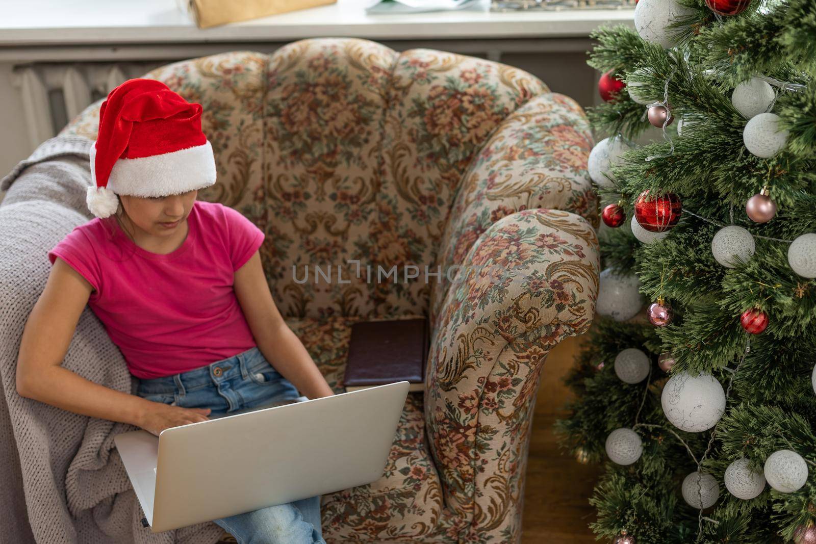 A happy little girl opens a gift under the Christmas tree at home and communicates with her grandparents via a laptop via video link. Holiday concept for Christmas and New Year at home.