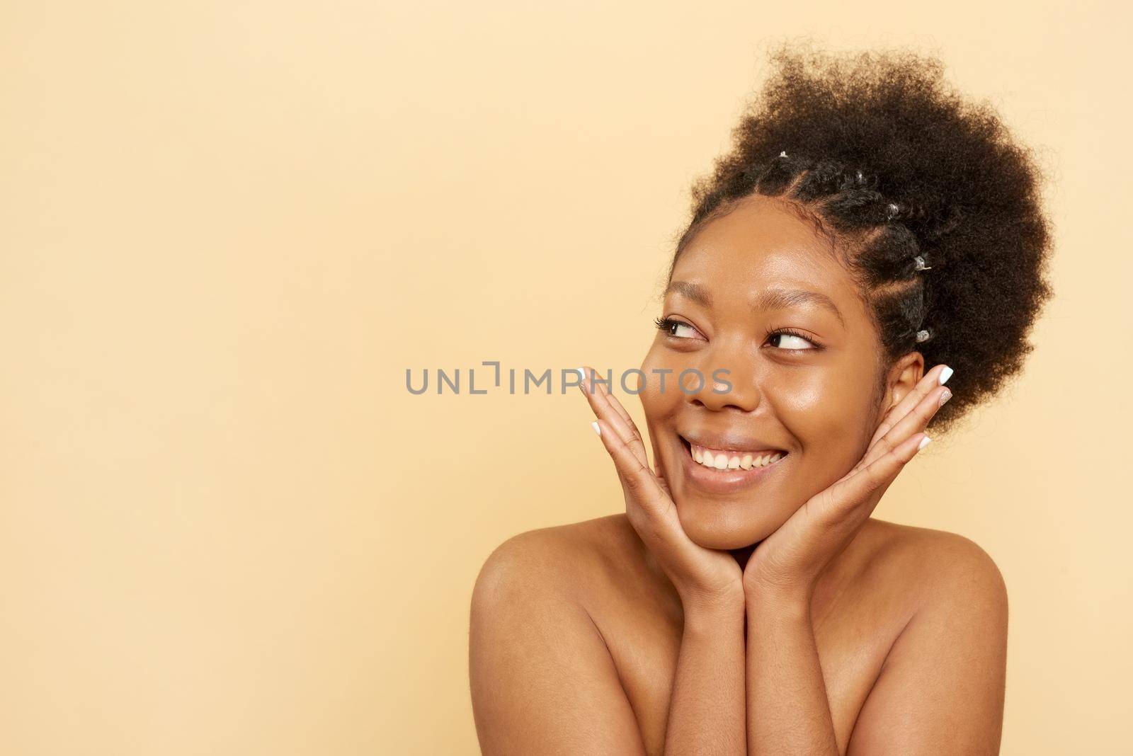 A beautiful young African American woman touches the delicate skin of her face. Happy beauty african lady after spa treatment isolated on beige with copy space. Beauty concept. by etonastenka